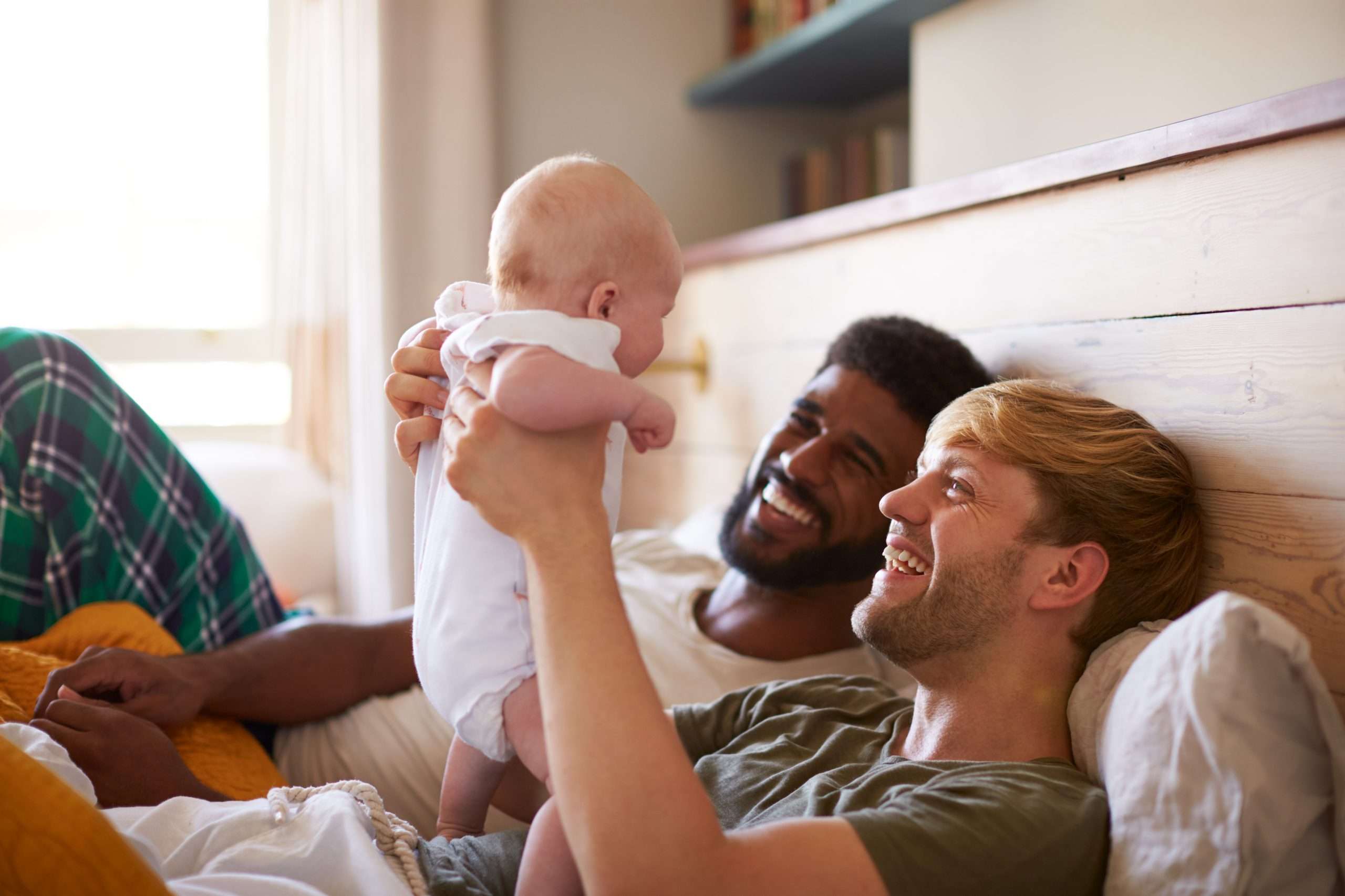 two men lay in bed smiling at baby